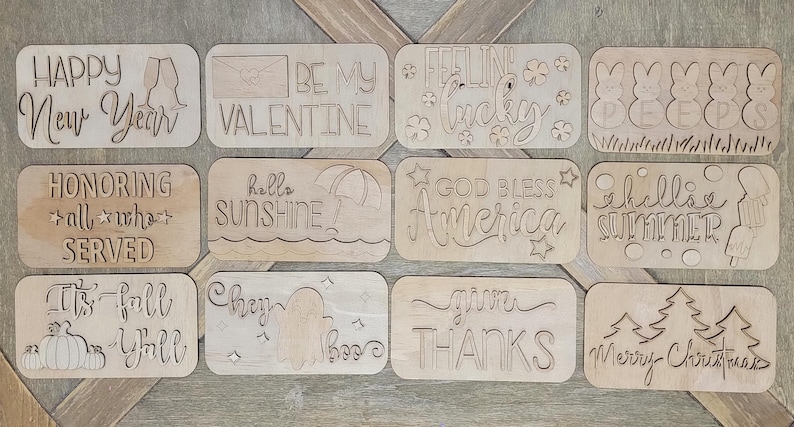 May include: A set of 10 wooden signs with various sayings, including "Happy New Year", "Be My Valentine", "Feeling Lucky", "Honoring All Who Served", "Hello Sunshine", "God Bless America", "It's Fall Y'all", "Cheery Boo", "Give Thanks", and "Merry Christmas".