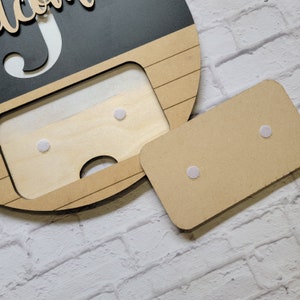 May include: A wooden sign with a cutout in the shape of a face. The sign is painted black with white lettering that says "Welcome". The cutout is filled with a piece of wood that has four white adhesive dots.