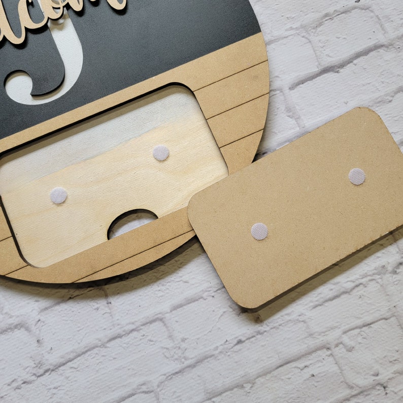 May include: A wooden sign with a cutout in the shape of a face. The sign is painted black with white lettering that says "Welcome". The cutout is filled with a piece of wood that has four white adhesive dots.
