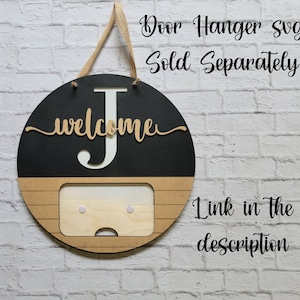May include: A black and brown wooden door hanger with the letter "J" and the word "welcome" in gold lettering. The hanger has a rectangular cutout in the bottom half, with two white dots on either side. The text "Door Hanger Svg Sold Separately Link in the description" is written in black text on a white brick background.