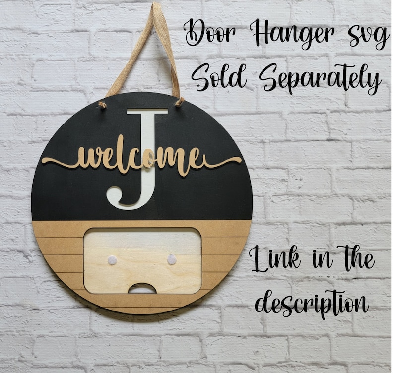 May include: A black and brown wooden door hanger with the letter "J" and the word "welcome" in gold lettering. The hanger has a rectangular cutout in the bottom half, with two white dots on either side. The text "Door Hanger Svg Sold Separately Link in the description" is written in black text on a white brick background.
