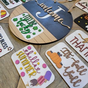 May include: A collection of wooden signs featuring seasonal greetings and sayings. The signs are decorated with various colours, patterns, and shapes. The signs include "Welcome", "Feeling Lucky", "It's Fall Y'all", "Happy New Year", "Be My Valentine", "Honoring All Who Served", "Merry Christmas", "Chillin' Summer", and "J's".