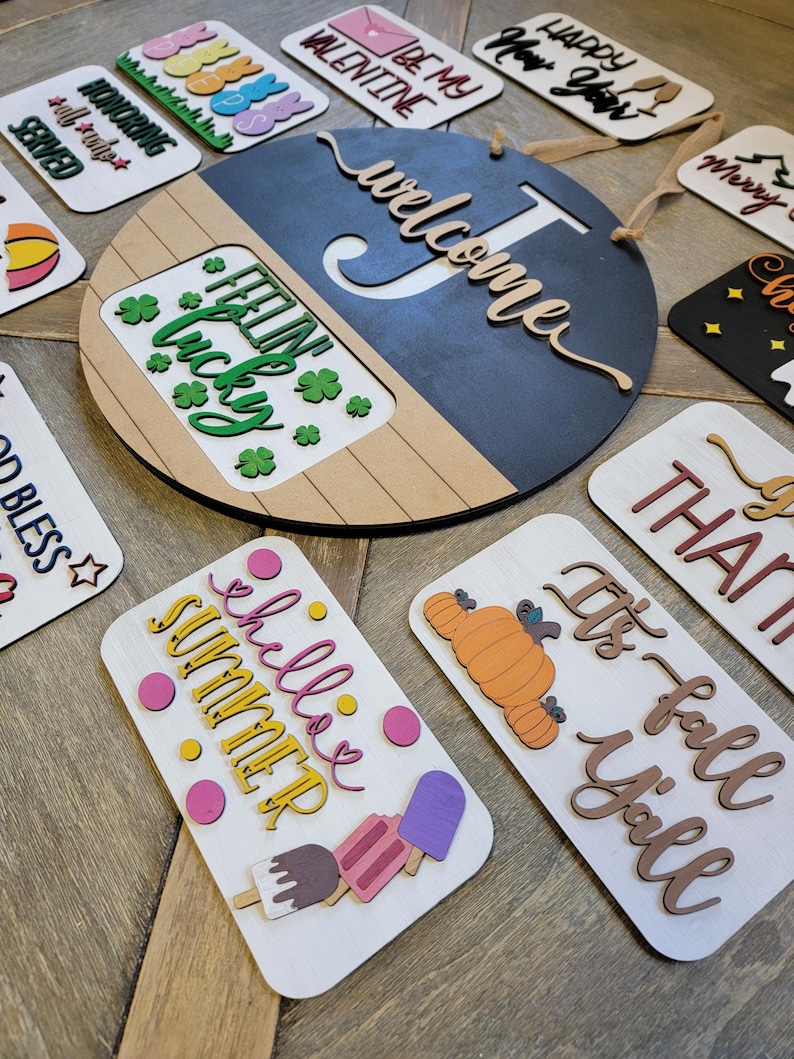 May include: A collection of wooden signs featuring seasonal greetings and sayings. The signs are decorated with various colours, patterns, and shapes. The signs include "Welcome", "Feeling Lucky", "It's Fall Y'all", "Happy New Year", "Be My Valentine", "Honoring All Who Served", "Merry Christmas", "Chillin' Summer", and "J's".