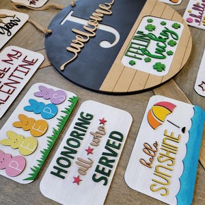 May include: A collection of wooden signs with various sayings and designs. The signs feature different colours, fonts, and styles, including a round sign with the word "Welcome" and a smaller sign with the text "Feeling Lucky".