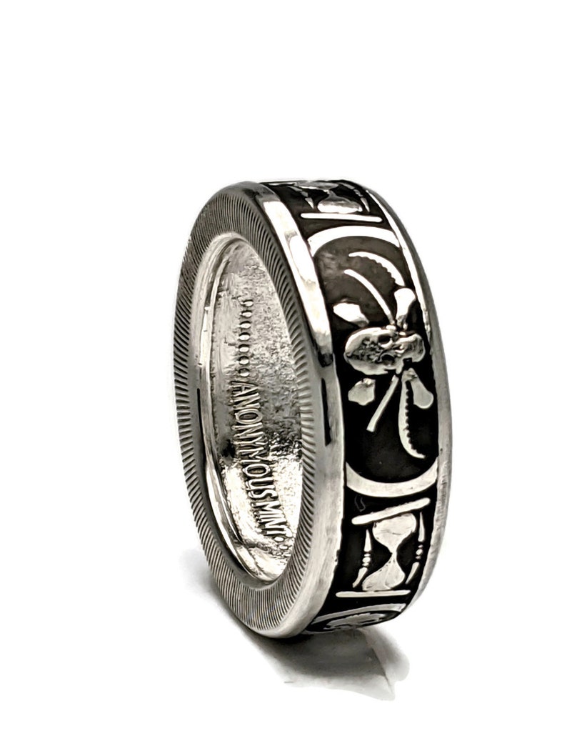 May include: A silver ring with a black design featuring an hourglass, a clover, and a skull. The ring is inscribed with "ANONYMOUS INC."