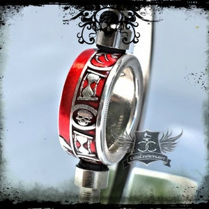 May include: A silver coin ring with a red enamel band. The band features a skull and hourglass design. The ring is on a stand and the text "Coincrafters.net" is visible.