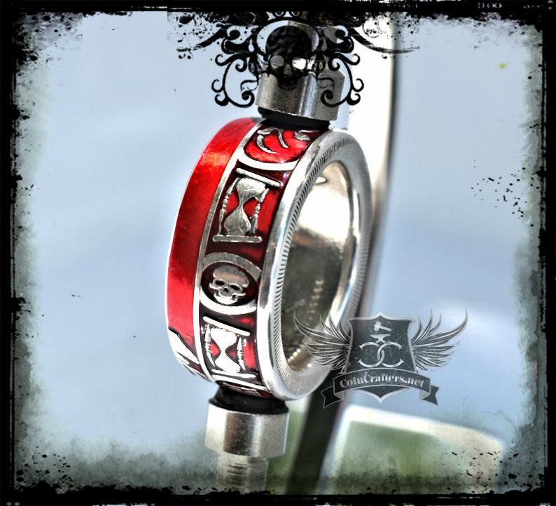 May include: A silver coin ring with a red enamel band. The band features a skull and hourglass design. The ring is on a stand and the text "Coincrafters.net" is visible.