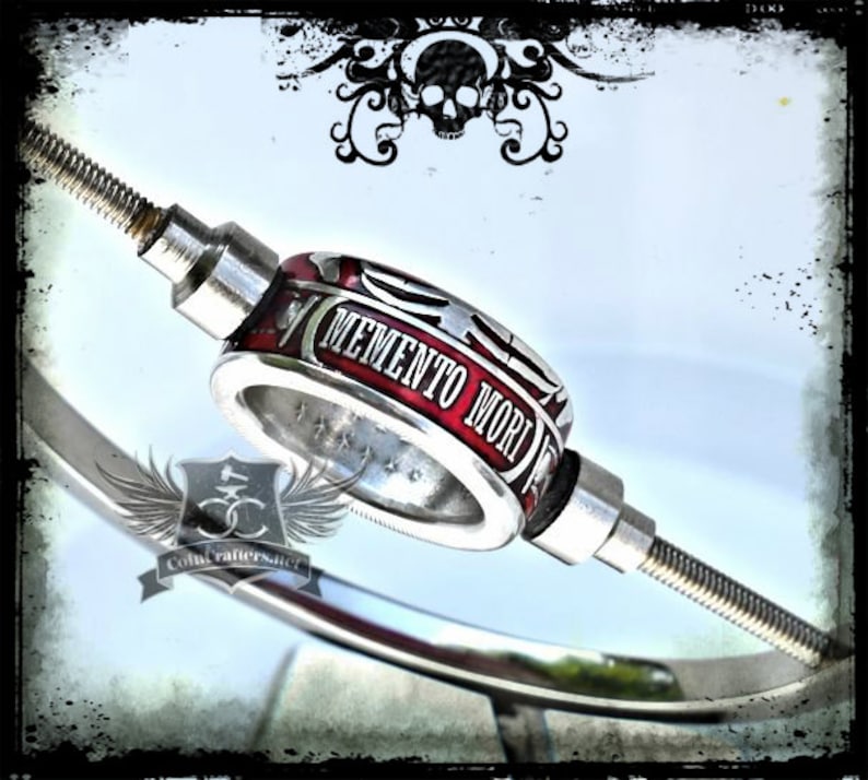 May include: A silver and red ring with the words "Memento Mori" engraved on it. The ring is attached to a silver rod.