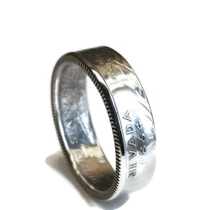 May include: A silver ring made from a coin, featuring the text "NEVADA 1864" and "UNITED STATES OF AMERICA". The ring has a detailed edge and a shiny, reflective surface. The ring is set against a white background.