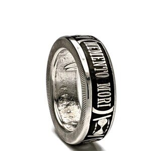 May include: A silver ring with a black band that says "Memento Mori" in white lettering. The band also features an hourglass design.