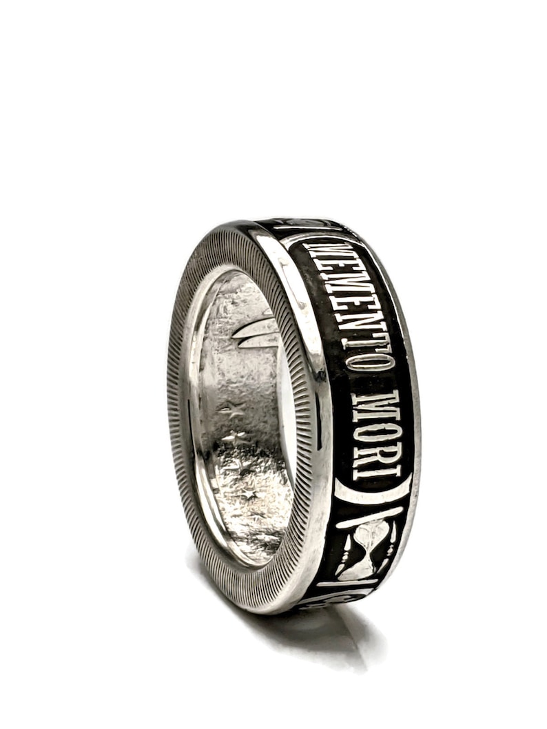 May include: A silver ring with a black band that says "Memento Mori" in white lettering. The band also features an hourglass design.