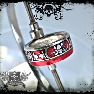 May include: A silver coin ring with a red enamel band and a white skull and crossbones design. The ring is on a metal stand.