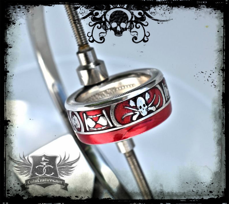 May include: A silver coin ring with a red enamel band and a white skull and crossbones design. The ring is on a metal stand.
