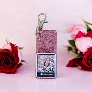 Peut inclure: Porte-clés en paillettes roses avec un anneau de clé argenté et une étiquette carrée comportant un code QR avec un visage de personne au centre. Le texte "s0lsky nn" est sous le code QR.