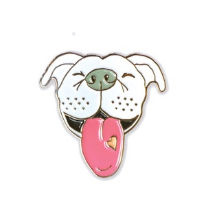 May include: White enamel pin of a dog with a pink tongue sticking out and a small heart on the tongue. The dog has a white face with black eyes and a green nose.