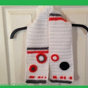 May include: White crocheted scarf with red, gray, and black details. The scarf features a design resembling a robot with circular shapes and a black circle.