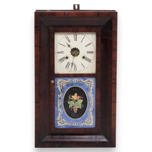 May include: Antique wooden wall clock with a rectangular frame. The clock face is white with black Roman numerals and hands. Below the clock face is a blue and black decorative panel with a floral design. The clock is a dark brown color.