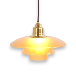 May include: A pendant light fixture with a layered, peach-colored shade and a brass-colored fixture. The light hangs from a dark brown cord against a white background. The light is turned on, emitting a warm glow.