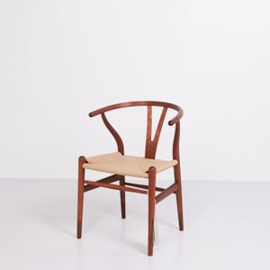 May include: A wooden Wishbone chair with a woven seat. The chair has a curved backrest and armrests. The wood is a warm brown color, and the seat is a light beige. A tag hangs from one of the legs.