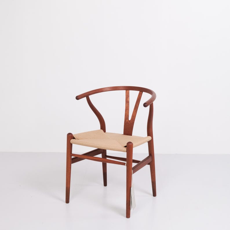 May include: A wooden Wishbone chair with a woven seat. The chair has a curved backrest and armrests. The wood is a warm brown color, and the seat is a light beige. A tag hangs from one of the legs.