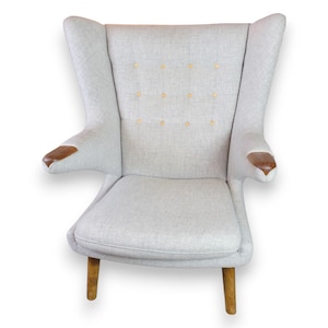 Peut inclure: Un fauteuil à dossier ailé rembourré gris clair avec accoudoirs et pieds en bois. La chaise a un dossier haut, des bras incurvés et un dossier capitonné. Les accoudoirs et les pieds en bois contrastent avec le tissu gris clair.