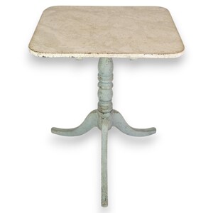 Op de afbeelding: Een kleine, vierkante, witgekalkte tafel met een lichtblauwe basis en drie poten. De tafel heeft een verweerde, vintage uitstraling.