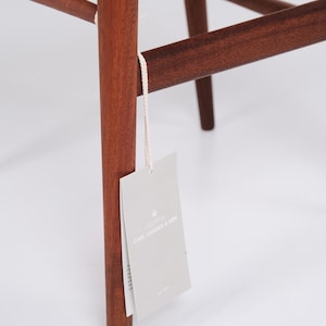 May include: Close-up of a wooden chair leg with a tag attached. The chair leg is a rich, reddish-brown color, and the tag is light gray with the text "CARL HANSEN & SON". The chair's design features clean lines and a minimalist aesthetic.