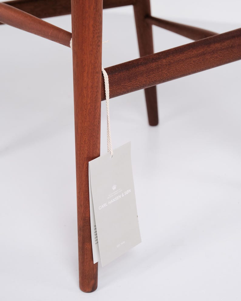 May include: Close-up of a wooden chair leg with a tag attached. The chair leg is a rich, reddish-brown color, and the tag is light gray with the text "CARL HANSEN & SON". The chair's design features clean lines and a minimalist aesthetic.
