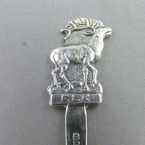 May include: A silver spoon featuring an elk figurine. The elk stands atop a base marked "ELK". The handle is engraved with "BRITISH COLUMBIA". The spoon is set against a grey backdrop. A souvenir spoon.
