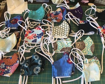 Può includere: Una collezione di maschere per il viso fatte a mano con vari motivi colorati, tra cui disegni floreali, geometrici e astratti. Le maschere sono realizzate in tessuto e hanno lacci elastici bianchi per le orecchie.