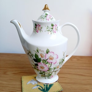 May include: A white porcelain teapot decorated with pink and white floral patterns and green foliage. The teapot has gold detailing on the spout and handle, and a gold-topped lid. It rests on a yellow coaster.