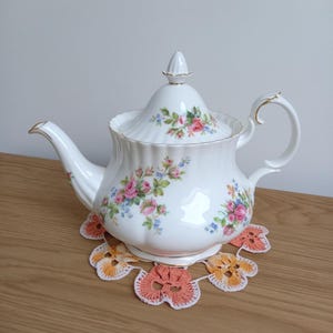 May include: A white porcelain teapot with a floral pattern of pink roses and blue flowers, gold trim. It rests on a crocheted doily with orange and white flower shapes. A traditional tea set, perfect for afternoon tea.