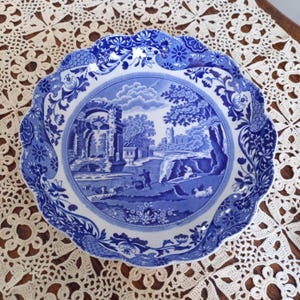 May include: A blue and white ceramic bowl with a scalloped edge. The bowl features a traditional blue and white transferware design of a landscape with a bridge, trees, and a building.