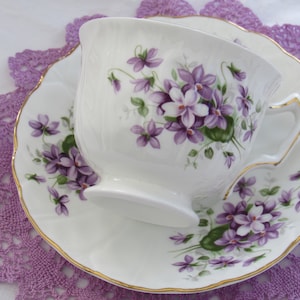 May include: A white teacup and saucer with a floral pattern of purple violets and green leaves. The cup and saucer have a gold trim.