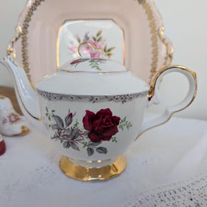 May include: A white porcelain teapot featuring a deep red rose floral pattern, a gold-trimmed base, and a matching lid. The teapot has a curved handle and spout, with a delicate grey floral border near the rim, set against a soft pink background.