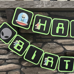 May include: Green and black alien-themed banner with the word "Happy" and the number "1" in a green font. The banner is strung together with a white string.