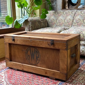 May include: A wooden storage trunk with a hinged lid and metal hardware. The trunk is stained a dark brown colour and has a circular logo with the letters "GWR" in the centre. The trunk is sitting on a patterned rug.