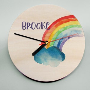May include: A wooden clock featuring a rainbow and cloud design. The clock face is white with black clock hands and the name "BROOKE" written in purple.