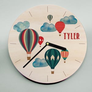 May include: A wooden clock with a light brown background featuring a design of hot air balloons in various colours, including red, blue, green, and yellow. The clock has black hands and a white face with black tick marks. The name "TYLER" is written in red letters at the bottom of the clock.