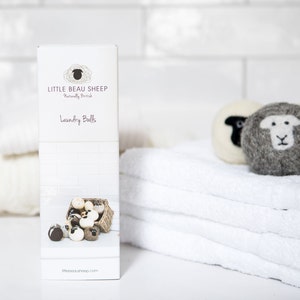 Puede incluir: Una caja blanca con el texto "Little Beau Sheep" y "Laundry Balls" en ella. La caja est&aacute; sobre una superficie blanca con una pila de toallas blancas y algunas bolas de lavado de lana con caras de ovejas.
