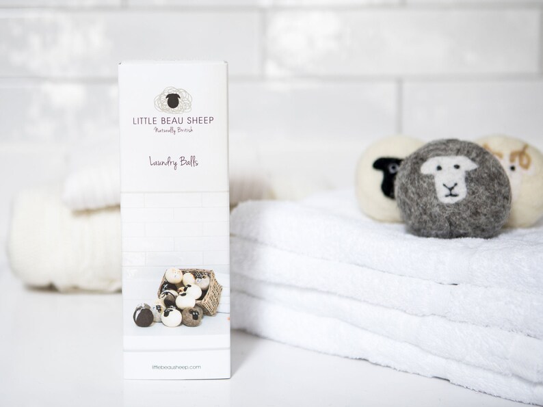 Puede incluir: Una caja blanca con el texto "Little Beau Sheep" y "Laundry Balls" en ella. La caja est&aacute; sobre una superficie blanca con una pila de toallas blancas y algunas bolas de lavado de lana con caras de ovejas.