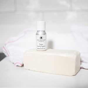 May include: A small silver spray bottle with a white cap sits atop a large, rectangular bar of soap. The bottle is labeled "Modern Laundry Fragrance" and "Little Beau Sheep." A white towel with pink trim is in the background.