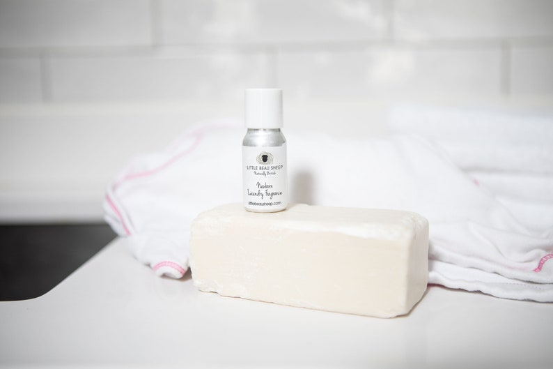 May include: A small silver spray bottle with a white cap sits atop a large, rectangular bar of soap. The bottle is labeled "Modern Laundry Fragrance" and "Little Beau Sheep." A white towel with pink trim is in the background.