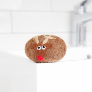 Puede incluir: Un jabón marrón en forma ovalada con un diseño de cara de reno. El reno tiene una cara marrón, cuernos blancos y una nariz roja. El jabón está sobre una superficie blanca, con una pared de azulejos blancos y un grifo cromado en el fondo.