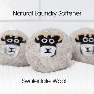 Puede incluir: Tres bolas de lana afieltrada de color gris claro, cada una con una cara de oveja hecha con fieltro de aguja en tonos negro, blanco y beige. El texto Natural Laundry Softener y Swaledale Wool está impreso en la imagen.