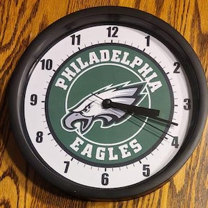 May include: Round black wall clock featuring the Philadelphia Eagles logo. The clock face is white with black numerals and hands. The team logo is centered, with a green background and a white and gray eagle.