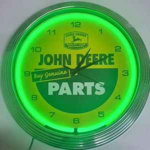 May include: A round, green and yellow John Deere clock with a neon green illuminated border. The clock face features the John Deere logo, the words "JOHN DEERE PARTS", and the phrase "Buy Genuine". The clock hands are black.