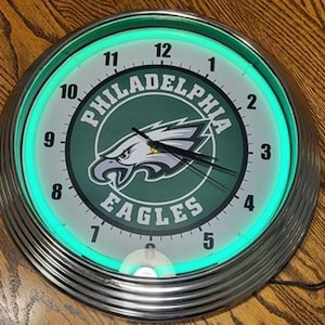May include: A neon green clock with a silver frame features the Philadelphia Eagles logo with the words "Philadelphia Eagles" in a green circle. The clock has black numbers and hands.