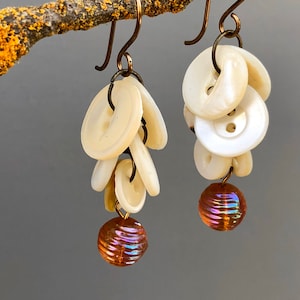 May include: A pair of dangle earrings featuring a cluster of white buttons and a single iridescent orange bead at the bottom of each earring.
