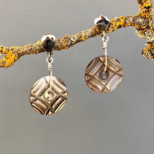May include: A pair of silver earrings with a brown, patterned button design. The earrings are hanging from a branch.